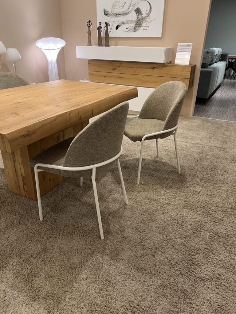 Mesa de comedor de madera con dos sillas tapizadas en gris claro y estructura blanca, junto a una lámpara de pie y un mueble flotante con decoración