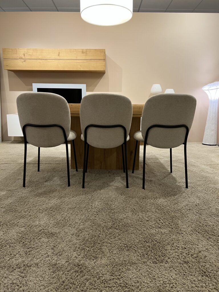 Tres sillas tapizadas en beige frente a una mesa de madera en una sala con alfombra beige y lámparas modernas