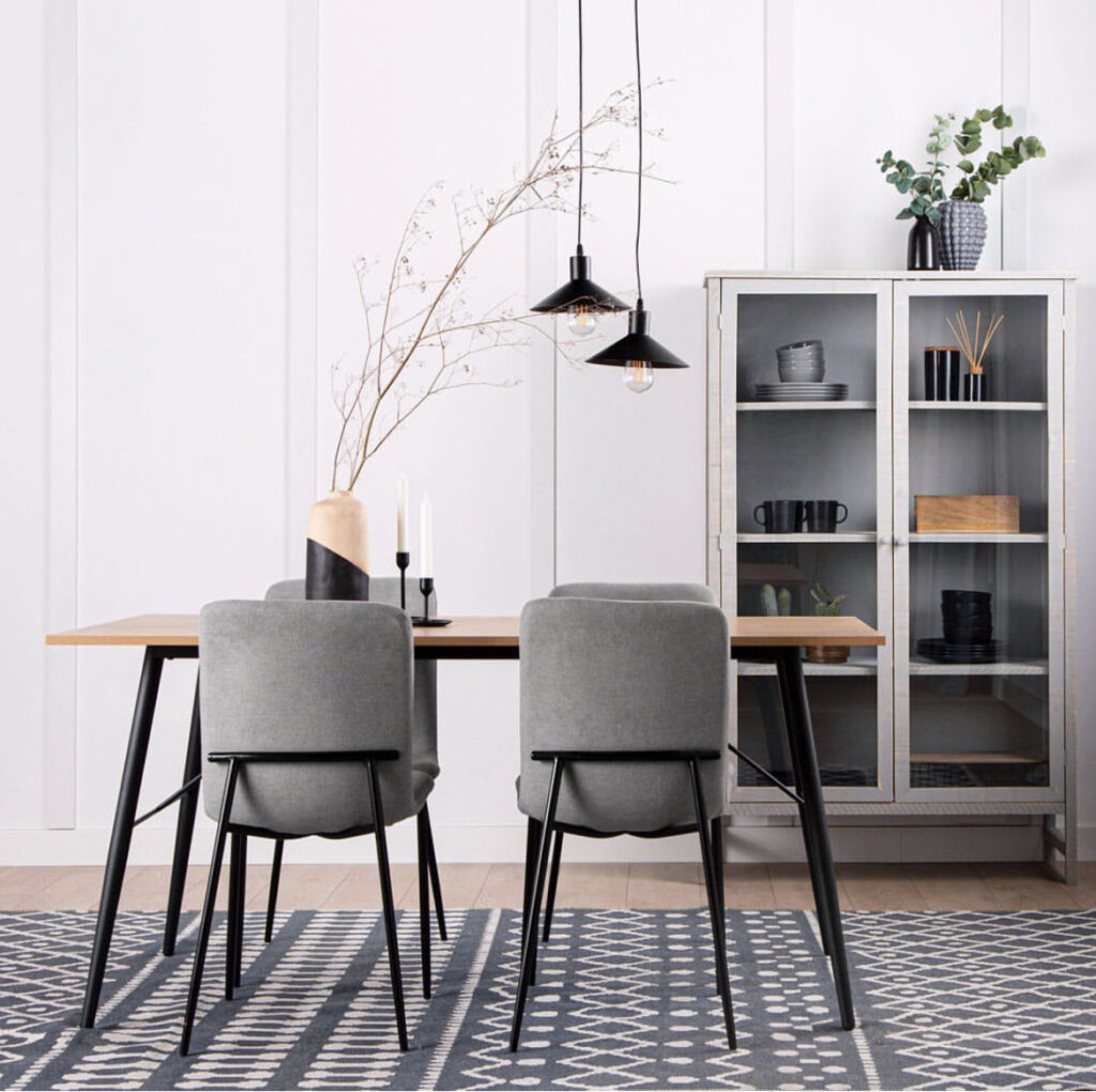 Comedor minimalista con mesa de madera, dos sillas grises, lámparas colgantes negras y vitrina con vajilla y decoración